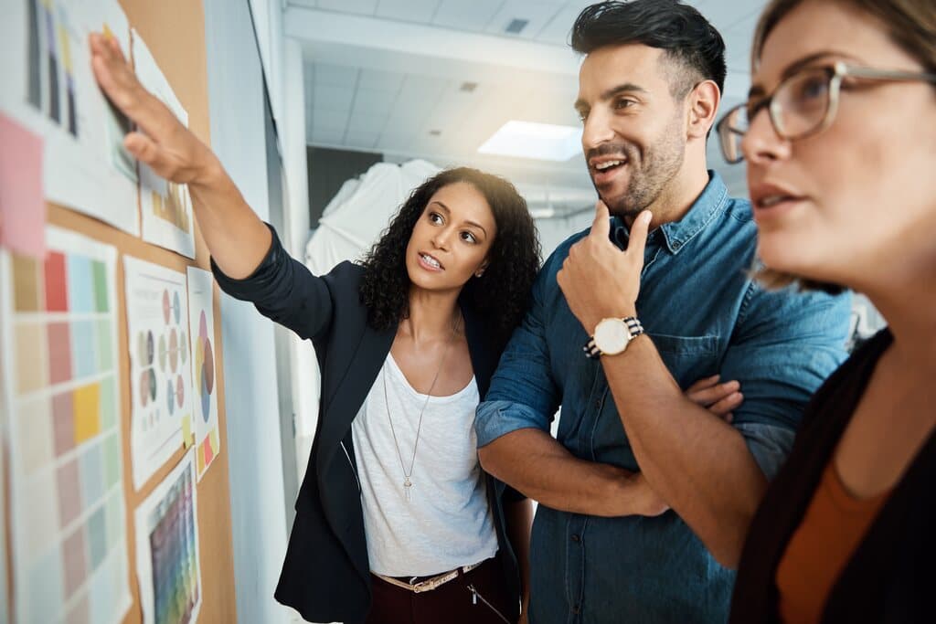 Três profissionais em um ambiente de escritório moderno analisam um quadro de cortiça com gráficos, paletas de cores e diagramas. Uma mulher aponta para um dos elementos enquanto explica sua visão aos colegas, demonstrando a aplicação prática de o que é Design Thinking através da síntese de informações e trabalho em equipe.
