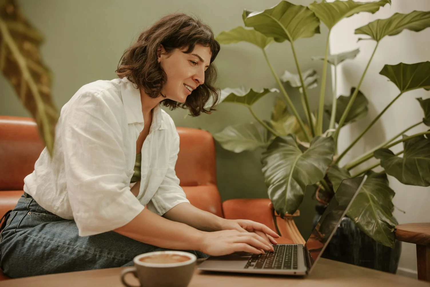 Mulher sentada no sofá de casa, sorrindo enquanto trabalha no notebook ao lado de uma caneca de café