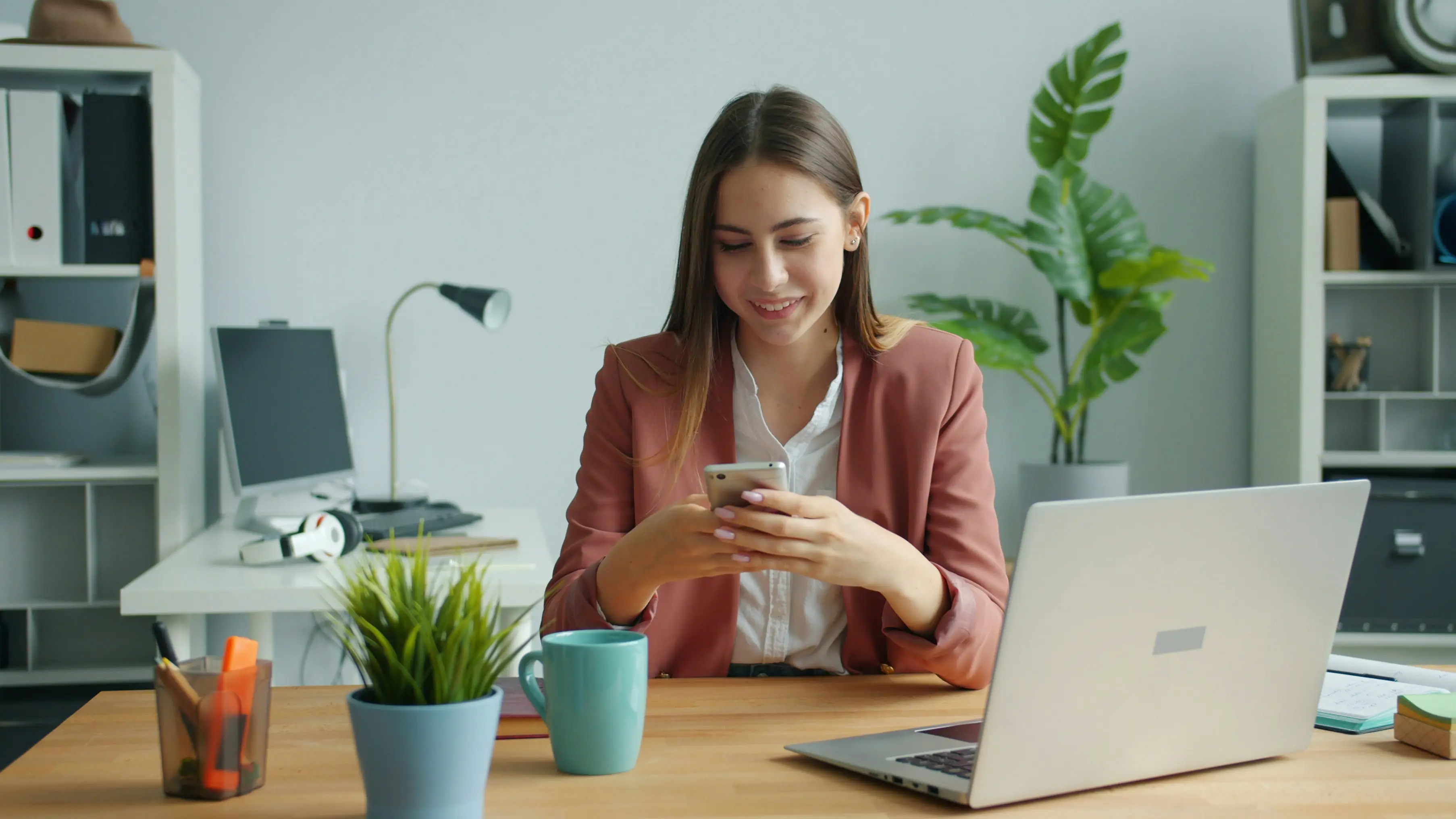 Mulher de blazer sentada à mesa de trabalho, sorrindo enquanto usa o celular ao lado do notebook
